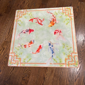 May include: Square decorative mat featuring a watercolor design of koi fish in shades of red, orange, and white, swimming on a light green background. The mat has a border with an orange geometric pattern and floral accents.