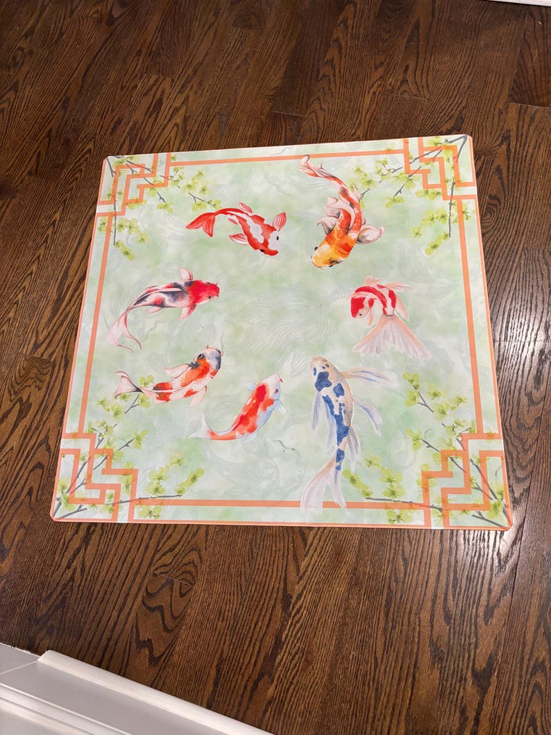 May include: Square decorative mat with a watercolour design of koi carp in red, orange, and white, swimming on a light green background. The mat has an orange geometric border and floral accents.