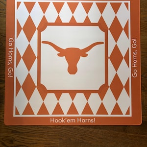 May include: A square orange and white mat featuring a central longhorn steer logo inside a white square. The border displays a repeating orange and white diamond pattern. The text "Hook'em Horns!" and "Go Horns, Go!" is printed on the edges.