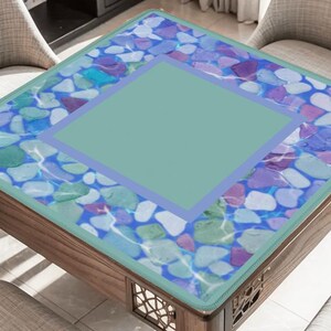 May include: A square wooden coffee table with a decorative glass top. The glass top features a design of colorful stones in shades of blue, purple, and green, with a central square in a solid light green color. The table is surrounded by two chairs.