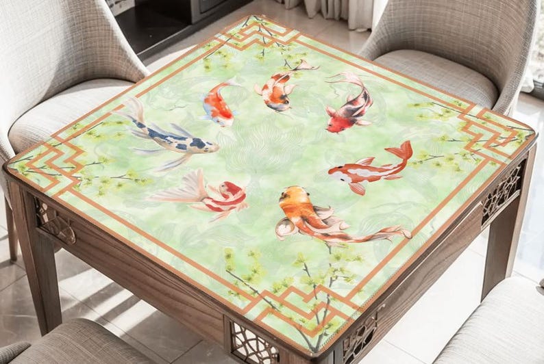 May include: A square wooden table with a decorative top featuring a watercolour design of koi carp in orange, red, and white against a green background. The table has a brown border and decorative cutouts.
