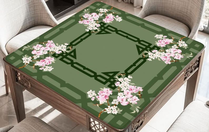 May include: A square table with a green floral design. The table's surface features a central geometric pattern and is adorned with pink and white blossoms. The table is made of wood and is surrounded by chairs.