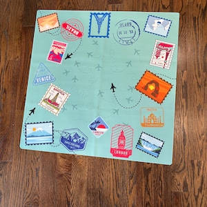 May include: A square light blue mat featuring a travel-themed pattern with various postage stamp illustrations of landmarks like the Eiffel Tower, Taj Mahal, and Big Ben, alongside travel stickers from Brazil, Venice, and Cancun with airplane icons.