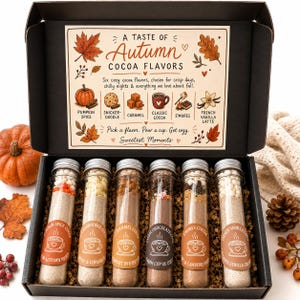 May include: A black gift box containing six glass test tubes filled with cocoa powder mixes. The box lid features a printed card titled A TASTE OF AUTUMN COCOA FLAVORS, listing six varieties: Pumpkin Spice, Snickerdoodle, Caramel, Classic Cocoa, S'mores, and French Vanilla Latte.