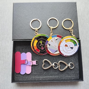 May include: A black gift box contains three keychains with colorful circular designs, a pink, purple, and magenta strap, and two heart-shaped clasps. The keychains have gold-colored rings. The circular designs feature a variety of colors.