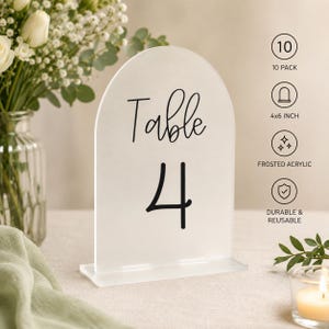 May include: An arched frosted acrylic table sign with the black text Table 4. The sign is part of a 10 pack, measures 4x6 inches, and is durable and reusable. The design features a smooth, semi-transparent surface with bold, dark lettering.