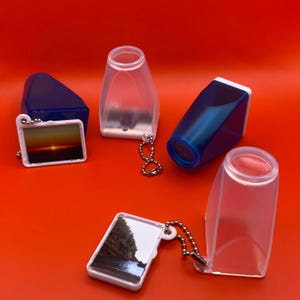 May include: An assortment of small, clear plastic magnifying glasses and keychain photo viewers are arranged on a bright orange background. The photo viewers display sunset and landscape images. The magnifying glasses are blue and clear.