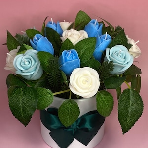 May include: A floral arrangement featuring blue, light blue, and white rose-shaped candles. The candles are arranged with green leaves and greenery in a white, round container with a dark green ribbon. The background is pink.