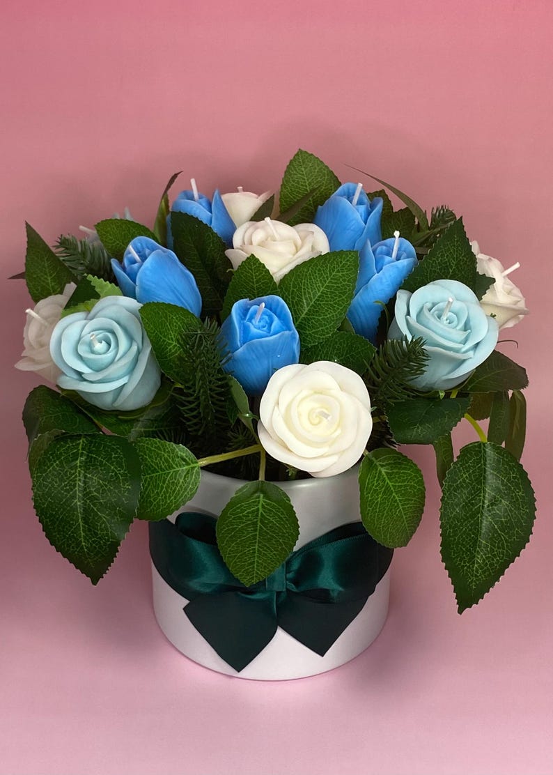 May include: A floral arrangement featuring blue, light blue, and white rose-shaped candles. The candles are arranged with green leaves and greenery in a white, round container with a dark green ribbon. The background is pink.