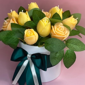 Puede incluir: Arreglo floral de velas amarillas en forma de rosa y hojas verdes en un recipiente blanco. Una cinta verde oscuro con una raya blanca adorna el recipiente. El fondo es rosa.