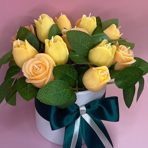 Puede incluir: Arreglo floral de velas amarillas en forma de tulipanes y rosas con hojas verdes en un recipiente redondo blanco. Un lazo verde oscuro con una cinta plateada adorna el recipiente.