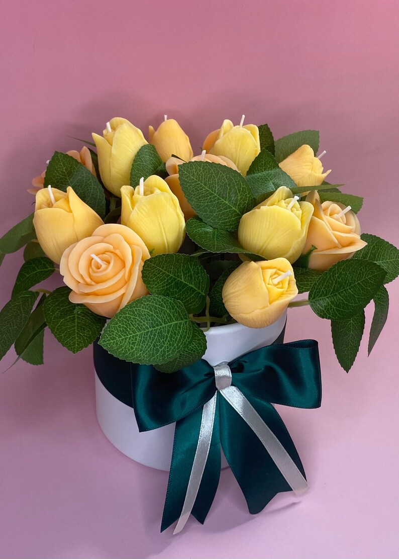 Puede incluir: Arreglo floral de velas amarillas en forma de tulipanes y rosas con hojas verdes en un recipiente redondo blanco. Un lazo verde oscuro con una cinta plateada adorna el recipiente.