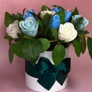 May include: A floral arrangement featuring white, light blue, and blue rose-shaped candles. The candles are nestled among green leaves and greenery inside a white round container. A dark green ribbon and bow adorn the container, set against a pink background.