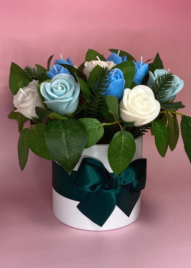 May include: A floral arrangement featuring white, light blue, and blue rose-shaped candles. The candles are nestled among green leaves and greenery inside a white round container. A dark green ribbon and bow adorn the container, set against a pink background.