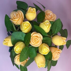 Puede incluir: Un ramo de velas con forma de rosas amarillas con hojas verdes. Las velas est&aacute;n en varias etapas de floraci&oacute;n, algunas abiertas, otras en forma de capullo. La disposici&oacute;n se presenta sobre un fondo morado claro, creando una exhibici&oacute;n visualmente atractiva.