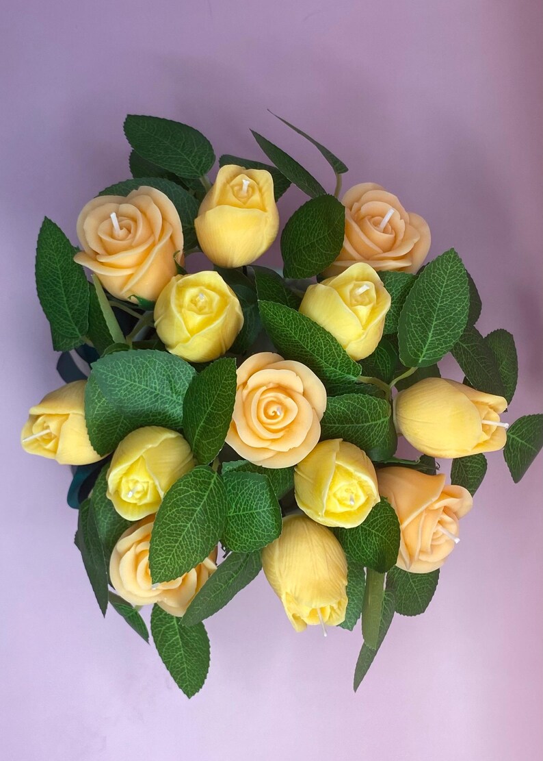 Puede incluir: Un ramo de velas con forma de rosas amarillas con hojas verdes. Las velas est&aacute;n en varias etapas de floraci&oacute;n, algunas abiertas, otras en forma de capullo. La disposici&oacute;n se presenta sobre un fondo morado claro, creando una exhibici&oacute;n visualmente atractiva.