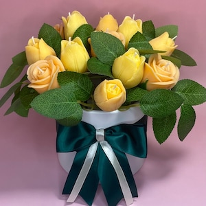 Puede incluir: Un arreglo floral de velas con forma de rosas y tulipanes amarillos con hojas verdes, presentado en una maceta blanca. La maceta est&aacute; adornada con un lazo verde oscuro con una cinta plateada. El fondo es rosa liso.