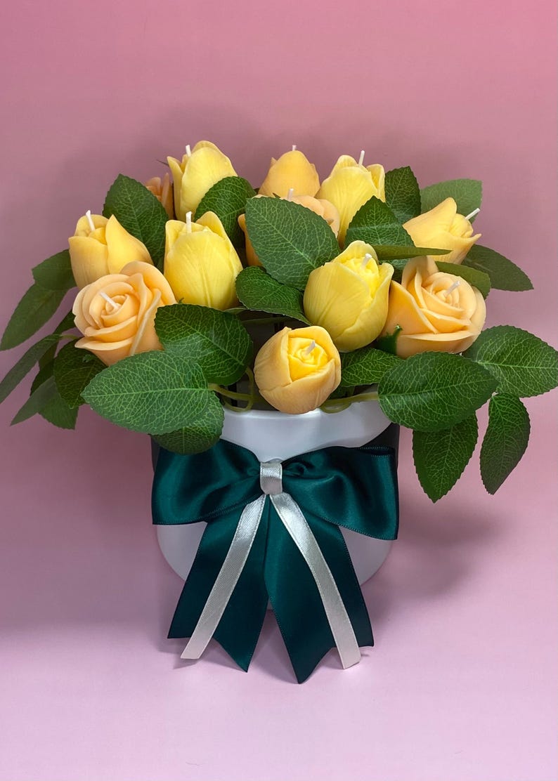 Puede incluir: Un arreglo floral de velas con forma de rosas y tulipanes amarillos con hojas verdes, presentado en una maceta blanca. La maceta est&aacute; adornada con un lazo verde oscuro con una cinta plateada. El fondo es rosa liso.