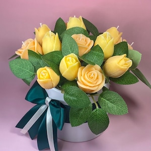 Puede incluir: Un ramo de velas amarillas con forma de rosa con hojas verdes en una maceta blanca. Una cinta verde oscuro con rayas blancas decora la maceta. El fondo es de color rosa liso.