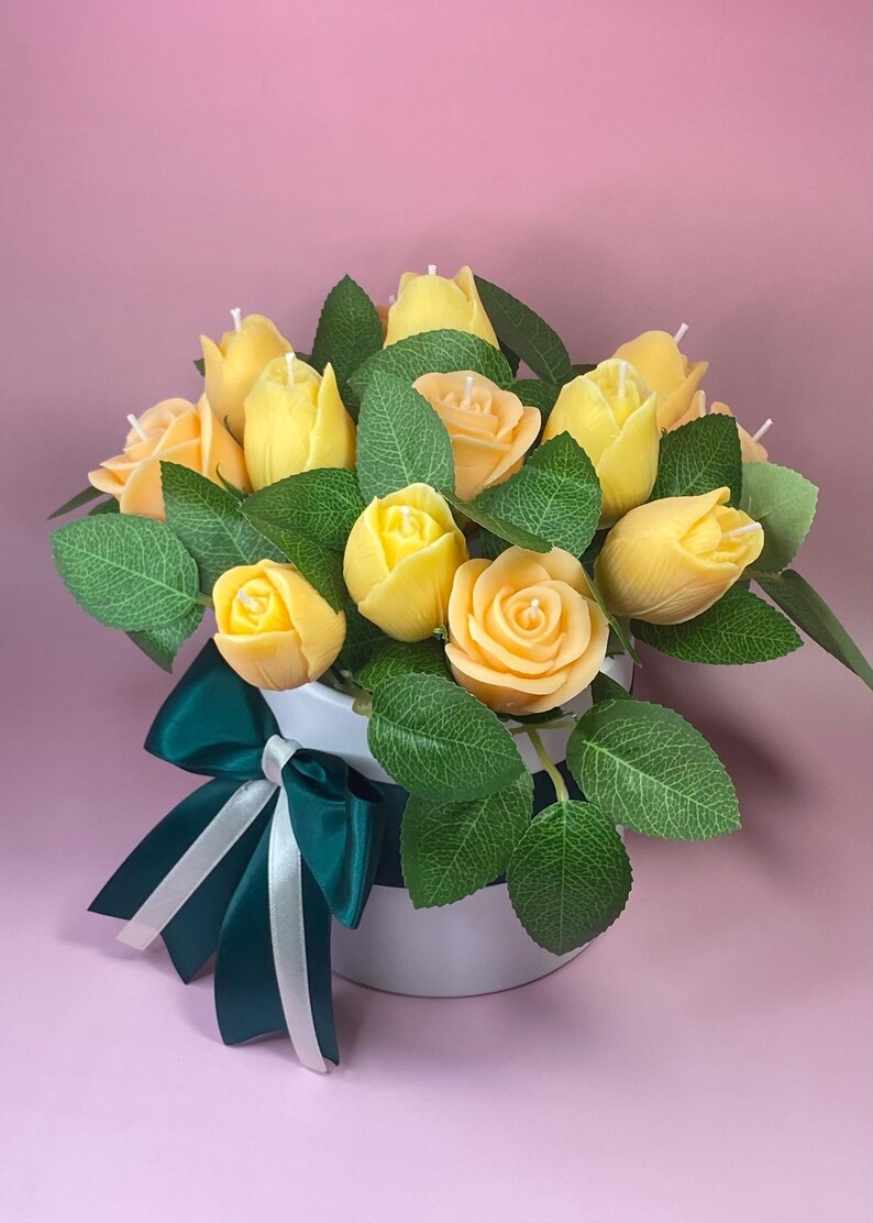 Puede incluir: Un ramo de velas amarillas con forma de rosa con hojas verdes en una maceta blanca. Una cinta verde oscuro con rayas blancas decora la maceta. El fondo es de color rosa liso.