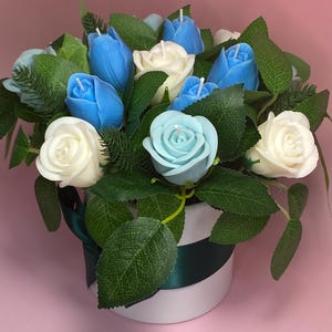 May include: A floral arrangement of candles in a white and green striped container. The arrangement features blue tulip candles, white rose candles, and a light blue rose candle, surrounded by green leaves and foliage. A dark green ribbon accents the container.