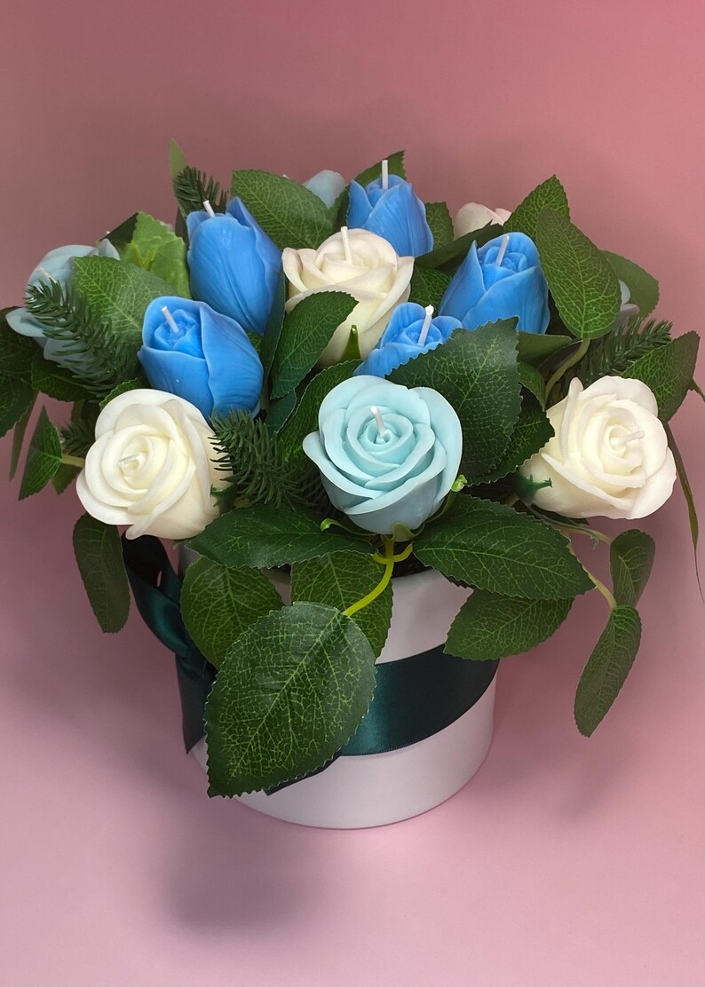 May include: A floral arrangement of candles in a white and green striped container. The arrangement features blue tulip candles, white rose candles, and a light blue rose candle, surrounded by green leaves and foliage. A dark green ribbon accents the container.