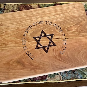 Könnte beinhalten: Ein rechteckiges Holzschneidebrett mit abgerundeten Ecken. Das Brett zeigt einen Davidstern in der Mitte, umgeben von hebräischen Texten. Die Holzmaserung ist sichtbar, und das Brett hat eine hellbraune Farbe.