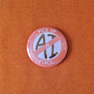 May include: A round pinback button with a peach-coloured background featuring a holographic star pattern. The text reads PICK UP A PENCIL, with the letters AI crossed out by a diagonal line. The design includes small colourful star icons.