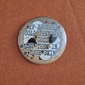 May include: A circular badge with the phrase "IF YOU'RE COLD THEY'RE COLD LET THEM IN YOUR SKIN" in white text. The badge has a brown background with black and white decorative elements.