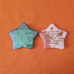 May include: Two star-shaped pinback buttons. The left button is dark green with light green text reading I SURVIVED THE TYEPOCALYPSE AND ALL I GOT WAS TRAUMATISED. The right button is light pink with brown text reading I survived a Mechanisms album and all i got was this dumb badge.