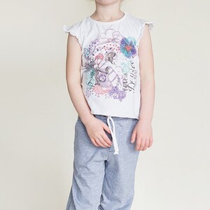 May include: A young girl in a white t-shirt with a colourful graphic print and grey sweatpants. The t-shirt has frilled sleeves and a drawstring waist. The girl is barefoot.