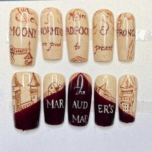 Puede incluir: Juego de diez uñas postizas con base crema y diseños en tinta marrón. Las uñas presentan texto e ilustraciones que recuerdan a un mapa, incluyendo las palabras "MOONY", "PRONG" y "The Marauder's Map". La parte inferior de algunas uñas es de color burdeos oscuro.