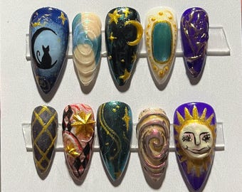 May include: A set of ten almond-shaped, press-on nails with various celestial and abstract designs. The nails feature a dark blue cat silhouette on a crescent moon, gold stars, and swirling patterns in shades of teal, gold, and purple.