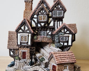 Może przedstawiać: Szczegółowa miniaturowa figurka domku z mostem, rzeką i małymi budynkami. Domek ma brązowy dach, białe i brązowe ściany oraz znak z napisem "The Potter's Mermaids".