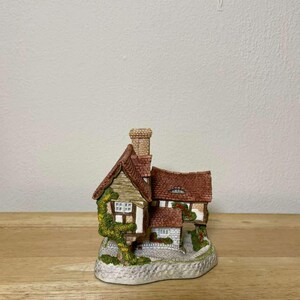 May include: A miniature cottage figurine with a red tile roof, brick chimney, and stone walls. The cottage has small windows and is adorned with green ivy. The base is a stone-like texture. A charming decorative piece.