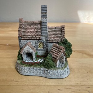 May include: A miniature stone cottage figurine featuring a tall gray chimney, a textured brown tiled roof, and a small arched entryway. The structure is accented with green foliage, a yellow-paned window, and a stone-textured base with white details.