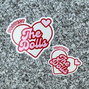 May include: Two stickers with a heart design and the text "Protect The Dolls". The stickers have a white background with pink text and a red outline. One sticker is larger than the other.