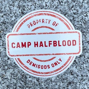 May include: A white and red circular sticker with the text "PROPERTY OF CAMP HALFBLOOD DEMIGODS ONLY". The sticker is on a speckled gray surface.