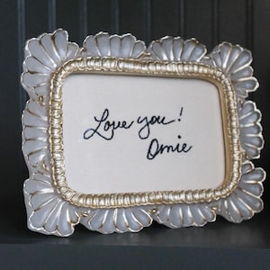 Pode incluir: Uma moldura retangular com acabamento dourado e uma borda branca e prateada com vieiras. A moldura contém um cartão branco com a mensagem manuscrita "Love you! Omie" em tinta preta.