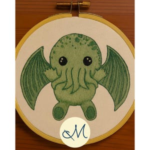 May include: An embroidered hoop art piece featuring a green Cthulhu monster with large wings and big black eyes. The embroidery is set in a yellow wooden hoop. A cursive "M" is at the bottom.