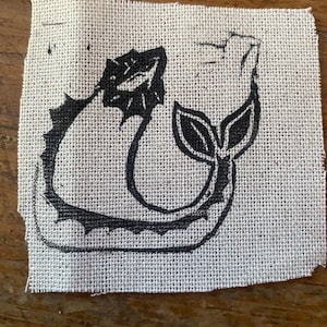 Może przedstawiać: Kwadratowy kawałek naturalnej tkaniny jutowej z czarnym nadrukiem linorytu stylizowanej syreny lub ryby. Projekt przedstawia zakrzywione ciało, ogon z dwoma liśćmi i dekoracyjną krawędź.