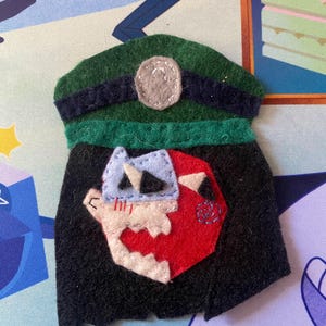 Op de afbeelding: Een vilten speld in de vorm van een groene hoed met een zwarte rand en een grijs embleem. Onder de hoed zit een zwarte cape met een wit, rood en blauw cartoon gezichtsontwerp. De speld is waarschijnlijk handgemaakt.