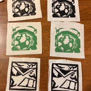 Puede incluir: Seis pequeños parches de tela cuadrados con diseños de bloques de impresión. Dos parches presentan un diseño de rostro en blanco y negro, dos tienen un diseño de rostro en verde y blanco y dos tienen un diseño de sobre en blanco y negro. Los parches están sobre una superficie de madera.