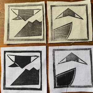May include: Four square fabric patches with black linocut prints. Each features a geometric design with a mountain, boat, and abstract shapes. The patches are on a wooden surface.