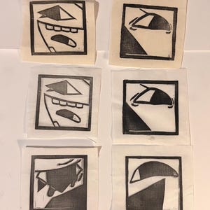 May include: A collection of nine small, square linocut prints on off-white fabric. The prints feature abstract, geometric designs in black ink, including stylized faces and shapes. The prints are arranged in a grid pattern.