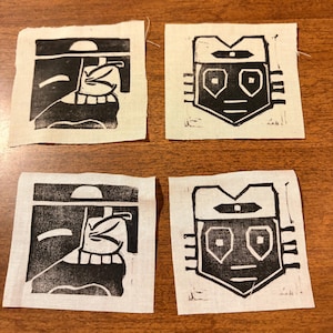 May include: Four small, square fabric patches with black linocut prints. Two patches feature a stylized face with a hat, and the other two depict a geometric face. The patches are on a wooden surface.