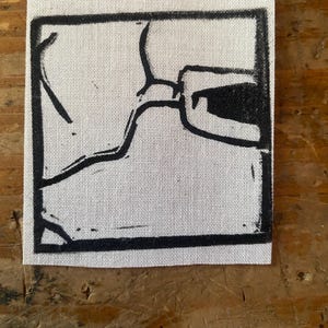 Może przedstawiać: Kwadratowa, biała naszywka z czarnym, abstrakcyjnym wzorem. Wzór to seria czarnych linii i kształtów w czarnej ramce. Tkanina ma widoczny splot.