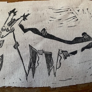 May include: A black and white linocut print on hessian. The artwork shows a stylized creature with a long body and a head with two antennae. The creature is holding flags and is next to a flower.