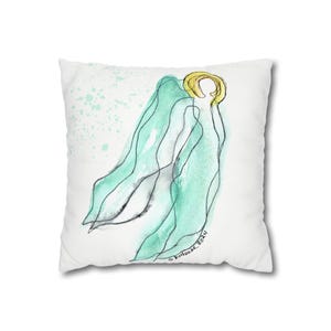 May include: White square pillow featuring a watercolor illustration of an angel. The artwork includes a gold halo, flowing teal-colored wings, and black outline details. The pillow has a soft, inviting appearance, perfect for home decor.