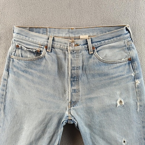 May include: Pale blue denim jeans featuring a button fly, traditional five-pocket layout, and distressed elements. The jeans display a faded appearance and signs of wear, including a small hole near the knee. A classic casual wear item.